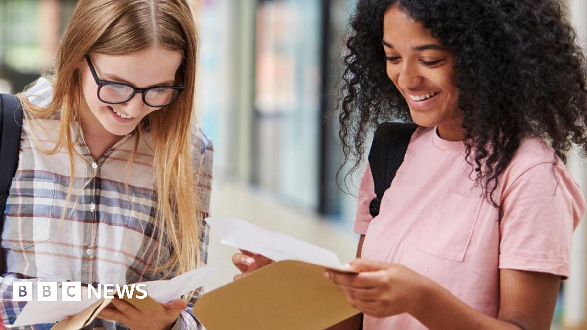 Two female students open exam results. The student on the elft is wearing a checkered shirt, has long straight blonde hair and wears black thick-rimmed glasses. The student on the right has dark curly hair and wears a pink t-shirt. Both students are smiling as they look down at the pieces of paper in their hands.