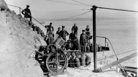 Mike Conner A black and white photograph showing a group of men in military uniform on a flight of stairs. A cannon can be seen in front of them, along with a group of metal containers. The sea can be seen behind them. 