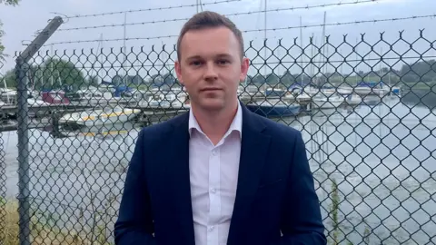 A man with short brown hair, wearing a navy suit and white shirt. There is a fence behind him. Behind the fence is Lough Neagh. 