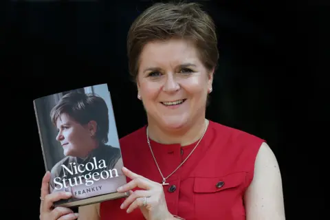 Getty Images Nicola Sturgeon, who  has short brown hair combed in a side shed and is wearing a sleeveless red blouse, holding a copy of her memoir, Frankly.