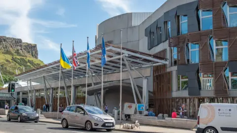 Getty Images The front of the holyrood building on a sunny day, with white stone walls wooden slatted windows and small trees and flagpoles with the scottish, UK and Ukraine flags