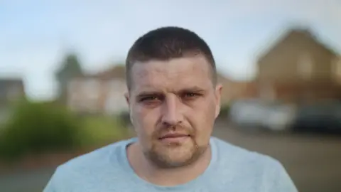 Ross Hart, a man with close-cropped brown hair and stubble, wearing a grey T-shirt, looking directly into the camera in a close-up shot with a shallow depth of field, so that the houses behind him are blurred