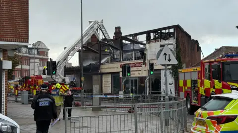 Elliot Deady/BBC Fire crews and police on the scene this morning. Hoses are being used to spray down a brick building with missing windows, a missing roof and charred walls. The sign on the front of the building is pale yellow and reads 'easy mobility services'.