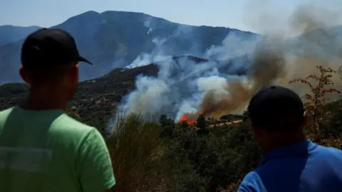 Reuters Two people both wearing hats have their backs to the camera as they look on as smoke rises from a wildfire burning in the distance with white smoke and orange flames fanning