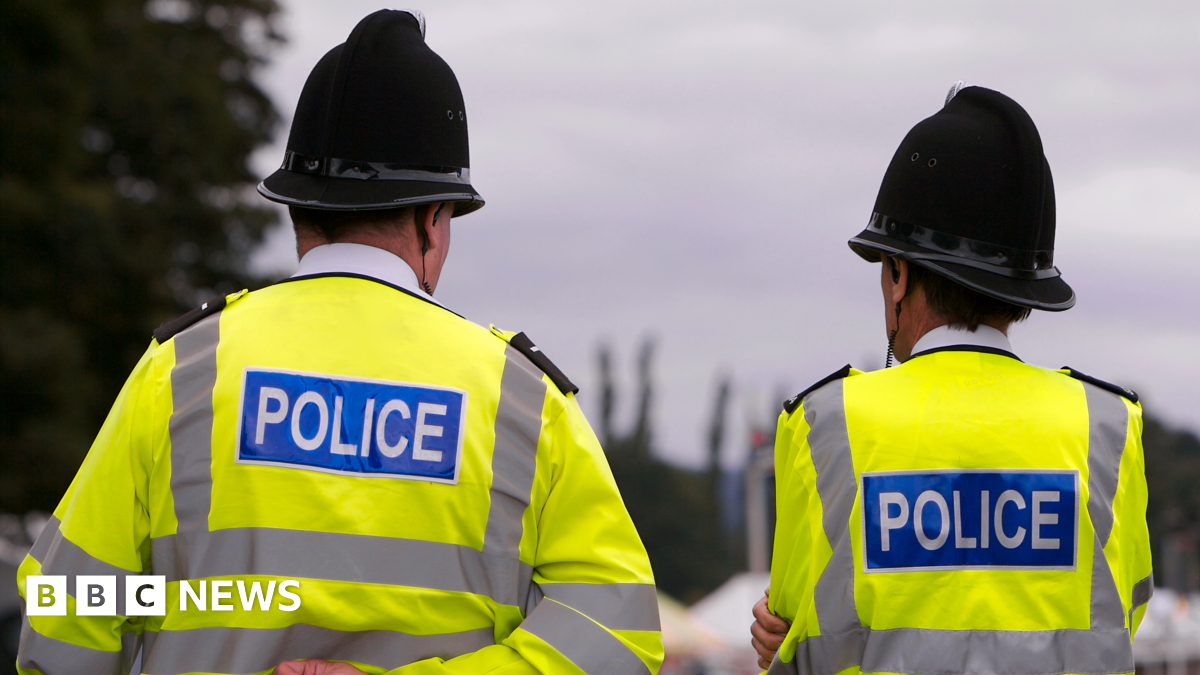 Two police officers stand with their backs to the camera, side by side in hi-vis jackets with 'POLICE' written on the back
