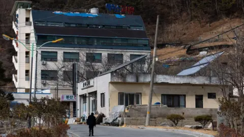Getty Images A person walks up a street towards a pair of blocky white buildings in front of a brown hillside, with trees growing on the side of the road