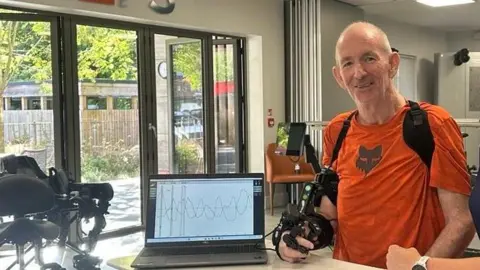 STEPS rehabilitation A man in an orange T-Shirt with a robotic device attached to his right arm, an open laptop next to him is displaying a series of wavelengths.
