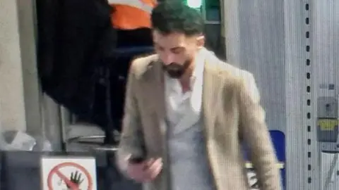 Essex Police A low-resolution CCTV still image of Ceyhan Dinler walking while looking at his phone. He is wearing a cream jacket and has a white shirt on.