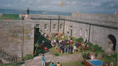 Mike Conner A group of people sit on benches in a courtyard, surrounded by large grey fortress walls. They are facing a band set up along the far wall. A small square plunge pool can be seen in the right hand corner. The  sea can be seen in the distance. 