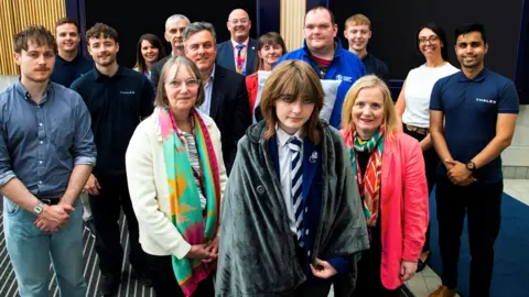Thales A schoolgirl in school uniform, with a heated blanket wrapped around her. Several other people are standing around her smiling for the camera. 