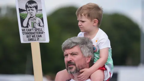 PA Media A man with grey hair and a grey beard has a young boy perched on his shoulders. The boy is holding a sign, which reads: "We can't spell 'cyanobacteria' - but we can smell it". The sign also depicts a young boy with two test tubes full of green fluid with a sad expression on his face. 