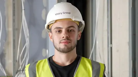 BBC Will Cunningham, a man in a white hard hat and hi-viz tabard, looks at the camera. He has a brown thin beard and moustache
