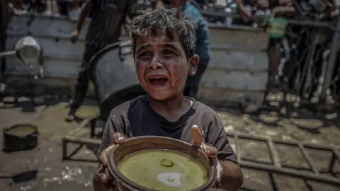 Anadolu via Getty Images A small crying boy, dirty with mud, holds a bowl up in search of food. Behind him more people are visible also looking for food. 