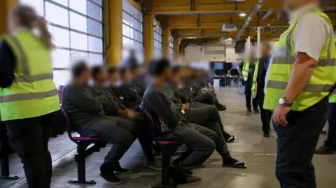 PA Media Staff in dark uniforms and hi-vis jackets look on as two rows of people in track suits and sandals sit waiting on two long rows of chairs in a large room that looks like a warehouse.     