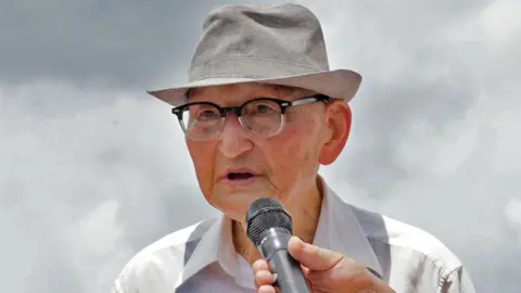 Jungmin Choi/BBC Ahn Hak-sop, wearing glasses and a hat, speaks with a mic being held close to his mouth