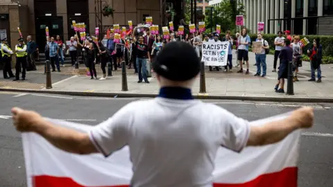 Jack Taylor/Getty Images A man holds a flag bearing the St George's cross opposite protesters attending a rally organised by Stand Up To Racism outside the Britannia International Hotel on July 25, 2025 in London, England. 