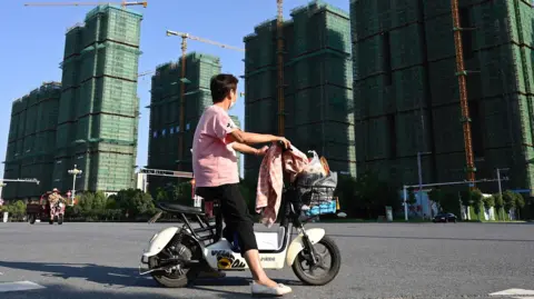 AFP via Getty Images A woman - wearing a mask, pink t-shirt with a Mickey Mouse emblem on the right sleeve and black trousers - rides a scooter past the construction site of an Evergrande housing complex in Zhumadian, central China's Henan province.