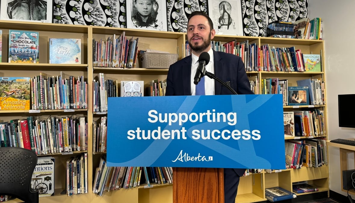 A man with dark hair and dark facial hair wearing a dark-coloured suit, white shirt, and blue tie stands behind a podium that reads "supporting student success." Behind him are brown wooden shelves filled with books.