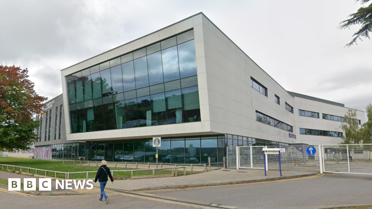 A college building with a large glass front and grey walls. A person is walking in front of the building.