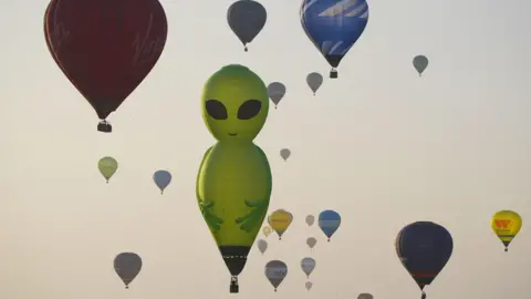 PA Dozens of balloons of all shapes and colours rise into the pale, clear morning sky at sunrise. The main focal point is the large, green balloon in the shape of an alien with black eyes.
