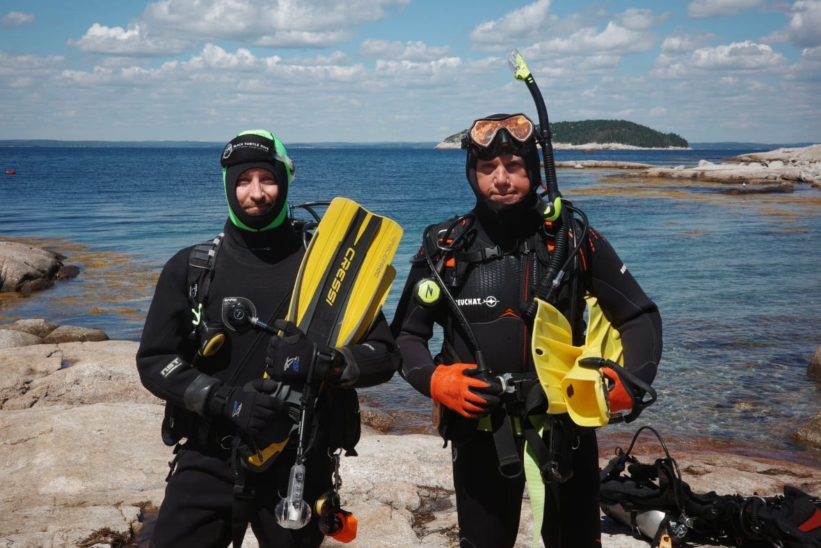 Two men in full scuba gear stand in front of the water. They're both wearing black wetsuits.
