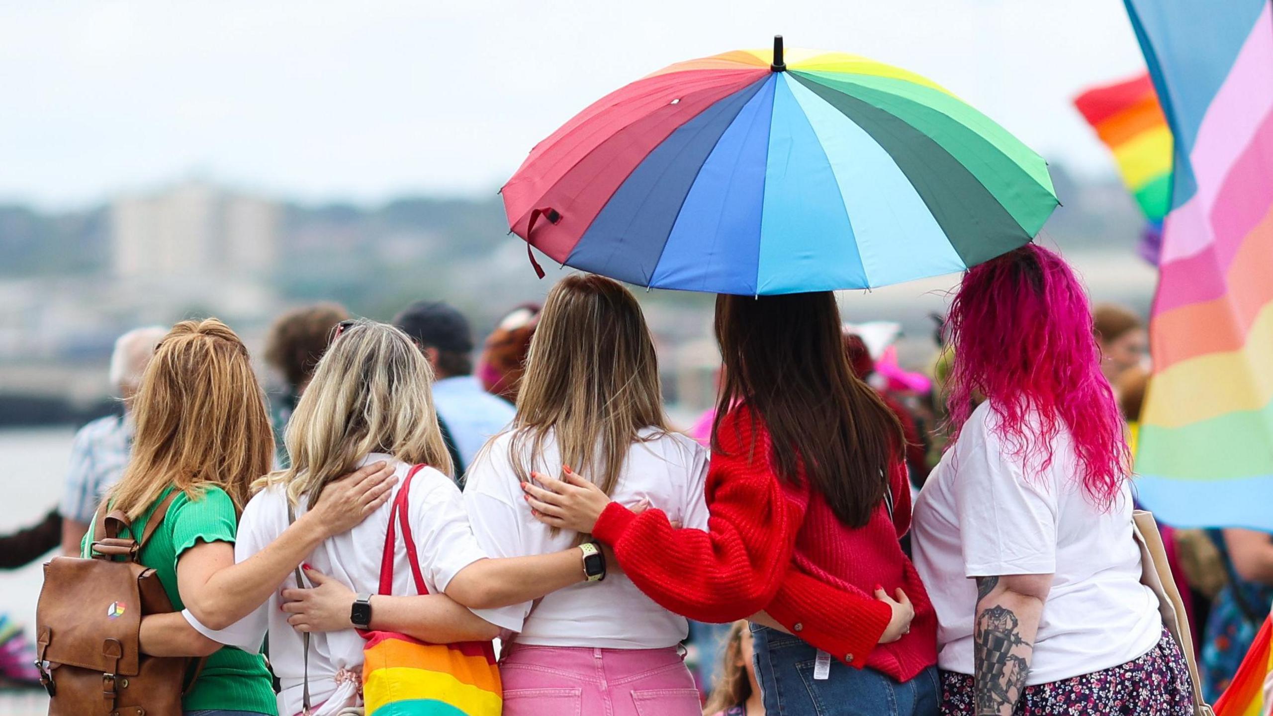 A group of people at Liverpool Pride are facing away from the camera and standing under a rainbow umbrella. They are wearing colourful clothes, and one has a rainbow bag while another has pink hair. A rainbow flag is nearby.