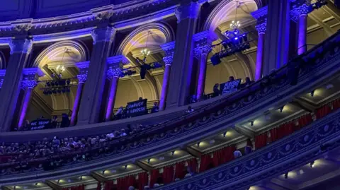 Supplied People standing in a concert hall below seated audience members hold up black signs with white writing on them