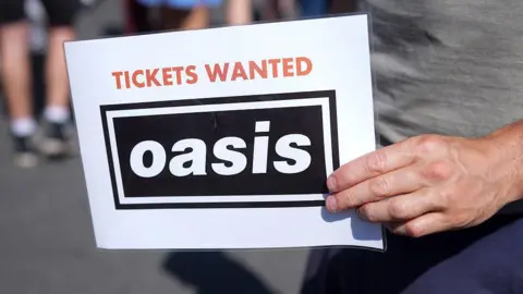 Getty Images A mans hand holds a piece of laminated white paper which reads 'Tickets wanted' in red font and a black box with white righting that reads 'Oasis.' The man holding the sign has on navy trousers and a grey top.