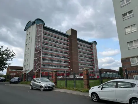 The block of flats in Leeds where Exodus died. Long balconies stretch between three main pillars, you can see cars parked outside and grass. 