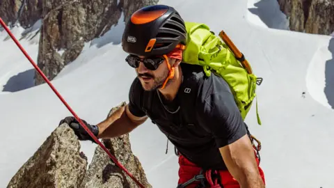 TOBY RONEY A climber wearing a black helmet, sunglasses, and a bright yellow backpack ascends a snowy mountain, gripping a rock, with snow-covered slopes and rocky cliffs in the background.