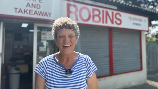 A woman with curly hair and wearing a blue striped t-shirt stands in front of a building with a red and white sign, which says Robins cafe and takeaway