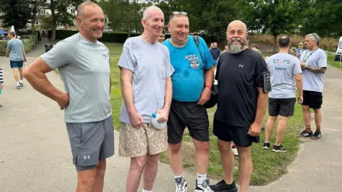 STEPS rehabilitation Four smiling men in shorts stood next to each other, looking past the camera at another photographer. Brendan is standing second from left wearing a grey T-shirt and shorts. There is grass and trees behind them.