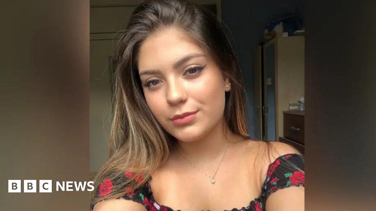 A young woman looks into the camera. She is wearing a black and red top and has long brown hair over her shoulders and brown eyes.