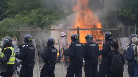 PA Media A line of officers with riot shields and helmets stand with their back to the camera, facing a large fire near some trees at the Holiday Inn Express in Rotherham