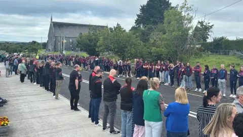 People lined up on a street outside a church. There is a cloudy sky above. 