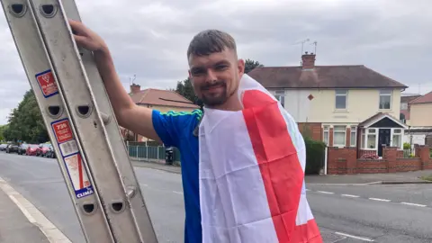 A man with a St George’s flag around his shoulders, wearing a blue polo shirt and holding onto a ladder on a road. He has short brown hair and a beard and is smiling at the camera, while cars and house can be seen behind him.