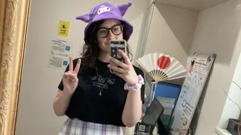 Al Thomann A person with shoulder-length wavy hair takes a selfie in a mirror, wearing a purple animal-shaped hat, a necklace and a black t-shirt. In the background is a computer and a fan with the Japanese flag