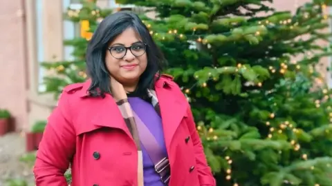 Sayanti Dutta A woman with dark shoulder-length hair and glasses in a bright pink coat stands next to a Christmas tree and smiles at the camera.