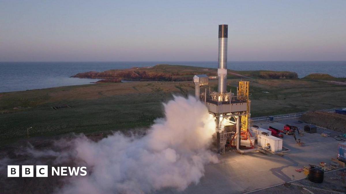A cylindirical silver rocket without a nose cone on the launch pad at a peninsula on an island in Shetland. White vapour is emerging from its thrusters as it prepares for launch. In the background is the sea.