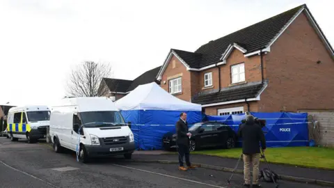 Getty Images A house with a blue forensic tent in front of it. The tent has a white roof. The house is red brick and has a dark-coloured roof with white guttering around it. In front of the tent is a white van. Behind the van is another van, but with police marking on it. In the foreground, a journalist wearing dark clothing and brown shoes stands in front of a camera with a cameraman wearing a dark jacket and light trousers.
