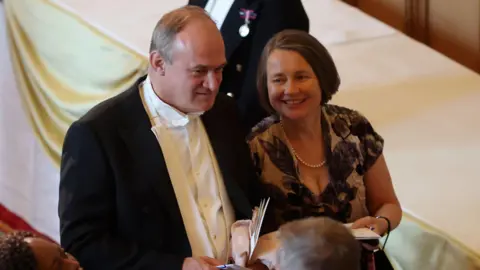 PA Media Liberal Democrat leader Sir Ed Davey walks alongside his wife Emily at the state banquet for Emmanuel Macron at Windsor Castle. He wears a formal black suit with white shirt and bow tie, while she wears a black and white floral dress with a pearl necklace.