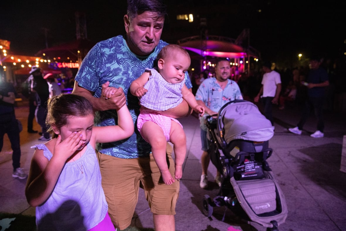 A man walks holding a baby in one arm and a young girls hand, another man behind them with a stroller