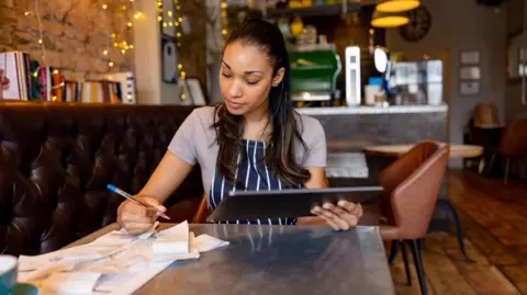 Getty Images A woman sitting in a bar restaurant wearing a striped apron and checking off a pile of receipts against a tablet