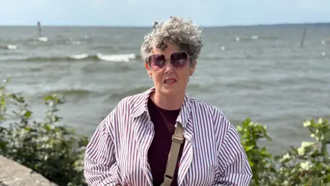 A woman is wearing a red striped shirt, cross body bag and sunglasses. She has grey curly hair and is stood in front of the shore of Lough Neagh