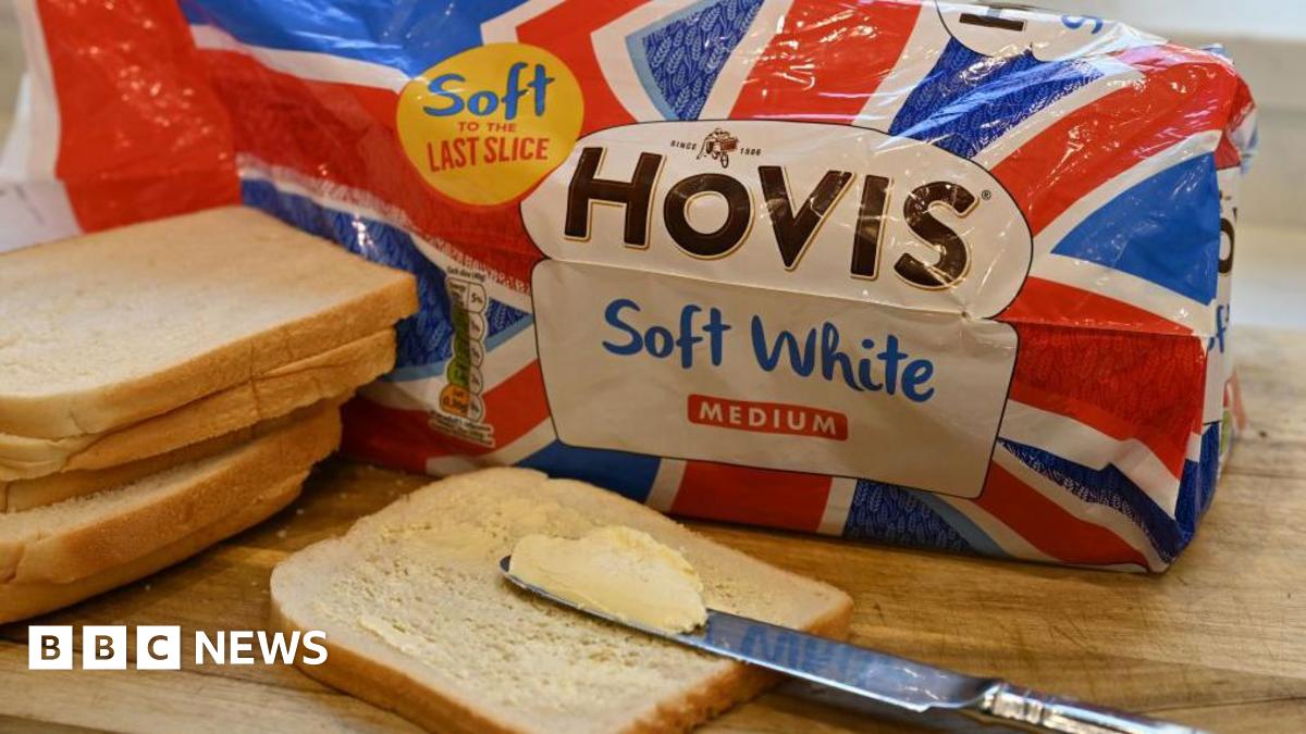 A loaf of Hovis bread in the packaging sitting on a bread board with slices piled around and a knife and butter on top of one slice in the foreground