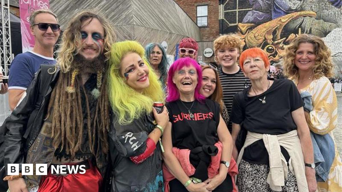 A group of people dressed in colourful outfits with colourful hair. They are standing on a street smiling at the camera. The walls behind them are covered in graffiti.