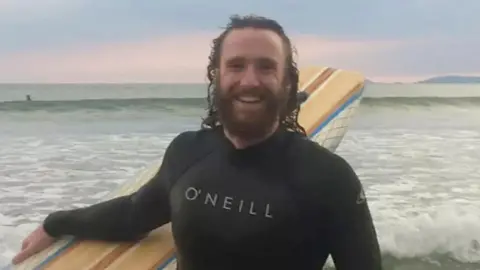 Family handout Alexander Barrett wearing a wet suit and holding a surf board while standing in front of the sea. He is smiling and has long brown hair and a brown beard.