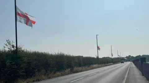 An A road which is lined with a hedge to the left, along every lamppost on the street is a St George's Cross flag