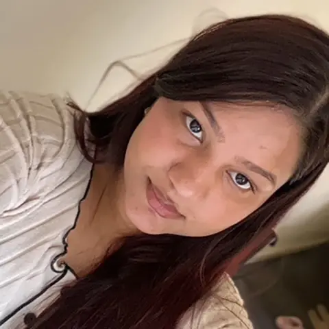 Nikita Upreti Nikita Upreti with long straight dark brown hair taking a selfie wearing a cream top with black piping and buttons with a neutral expression on her face