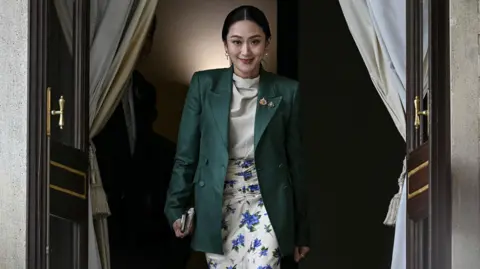 Getty Images Thailand's suspended prime minister Paetongtarn Shinawatra arrives for a press conference in Bangkok on July 1, 2025. She can be seen smiling as she walks through a doorway in a dark green blazer which she is wearing over a white shirt and white and blue floral skirt. 